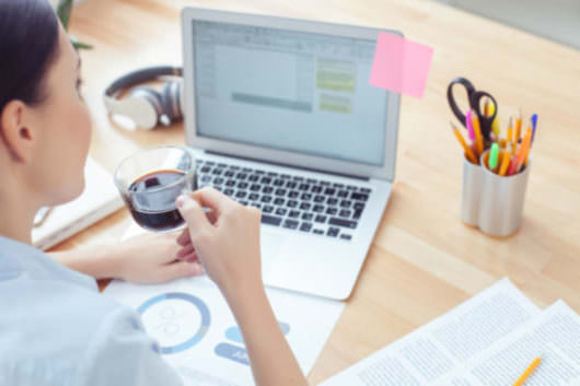 Pretty young woman is working with a laptop in office. She is drinking a cup of coffee with enjoyment. The lady is sitting at desk to learn Brazilian Portuguese.